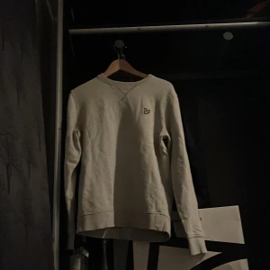 Ljusgrå sweatshirt från Lyle & Scott - Säljer en ljusgrå sweatshirt från Lyle & Scott med klassisk rund hals och broderad gul logga på bröstet. Tröjan har långa ärmar och ribbade muddar vid ärmslut och nederkant. Perfekt för en chill och stilren look.