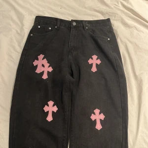 Chrome Hearts svarta jeans med rosa kors - Priset kan diskuteras! Unika svarta jeans från Chrome Hearts med flera rosa korsapplikationer på både fram- och baksida. Klassisk femficksmodell med silverfärgade nitar och detaljerad knapp. Jeansen har en loose passform och är tillverkade i robust denim.