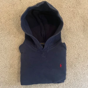 Marinblå hoodie från Polo Ralph Lauren - Säljer en marinblå hoodie från Polo Ralph Lauren med klassisk röd broderad logga på bröstet. Tröjan har huva, känguruficka och är tillverkad i mjuk bomull. Perfekt för chill dagar och snygg till jeans eller joggers.
