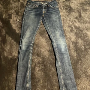 Blå skinny jeans från Nudie Jeans - Säljer ett par blå skinny jeans från Nudie Jeans med snygga slitningar och kontrastsömmar. Modellen har klassisk femficksdesign, orange detaljer och en läderpatch bak. Perfekt passform för dig som gillar smalare jeans.