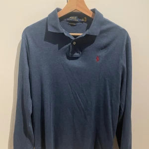 Blå långärmad pikétröja Polo Ralph Lauren - Snygg marinblå långärmad pikétröja från Polo Ralph Lauren i storlek S. Tröjan har klassisk krage, två knappar och rött broderat logotyp på bröstet. Perfekt för dig som gillar stilren och tidlös design.