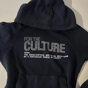 Svart hoodie med silvriga nitar - Mörkblå hoodie med texten 'FOR THE CULTURE' framtill, där bokstäverna är dekorerade med silvriga nitar för en extra cool vibe. Tröjan har huva och magficka, perfekt för dig som gillar streetstyle och vill sticka ut lite extra.