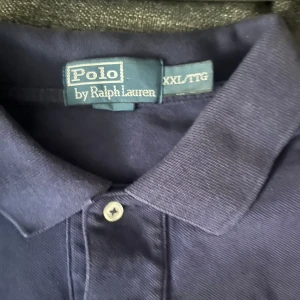 Marinblå pikétröja från Polo Ralph Lauren - Säljer en klassisk marinblå pikétröja från Polo Ralph Lauren i storlek XXL. Tröjan har krage, två knappar framtill och den ikoniska röda Polo-loggan broderad på bröstet. Tillverkad i mjuk bomull som är skön mot huden.