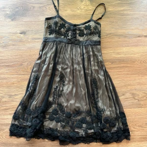 Svart meshklänning - Supersöt kortklänning från Indiska med tunna axelband och broderade svarta blommor på tyll. Klänningen har en beige bas, liten rosett framtill och vackra detaljer nertill. Perfekt för dig som gillar romantisk och unik stil.