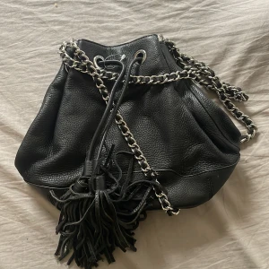Svart axelväska med kedja och fransar - Snygg svart axelväska från Rebecca Minkoff i skinn med grov silverkedja som axelrem. Väskan har snörning upptill och coola fransdetaljer framtill. Insidan är fodrad med svartvitt rutigt tyg och har praktiska fack. Perfekt för dig som gillar edgy stil.