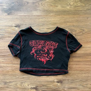 Svart crop top - Cool croppad svart topp från H&M med röd kontrastsöm och tryck med texten 'CHAOTIC' och en grafisk orm på bröstet. Rund halsringning och korta ärmar. Perfekt för dig som gillar edgy streetstyle och vill sticka ut med detaljerad print.
