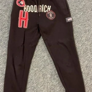 Svarta Hoodrich mjukisbyxor S - Svarta mjukisbyxor från Hoodrich med stora röda och vita tryck framtill, inklusive logga och patchdetaljer. Byxorna har snörning i midjan och smal passform. Perfekt för chill eller streetwear-stil.