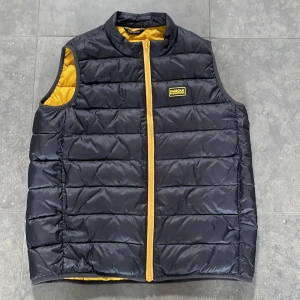 Svart dunväst från Barbour International - Svart quiltad dunväst från Barbour International med gul insida och gul dragkedja. Västen har ett Barbour International-märke på bröstet och är tillverkad i syntetmaterial med FIBREDOWN-fyllning. Perfekt för lager-på-lager och har en clean, sportig look. Skulle säga att den passar en lite mindre person typ en 11-12 åring som är typ 155-160