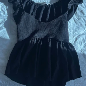 Svart volangtopp från 157 XS - Supersöt svart volangtopp från 157 i storlek XS. Toppen har volangdetaljer vid axlarna och en rynkad, flowy kjol. Materialet är lätt och har en snygg struktur som ger extra känsla. Perfekt för dig som gillar en feminin och trendig look med offshoulder-vibbar.