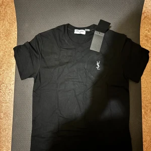T-shirt från Saint Laurent - Svart t-shirt från Saint Laurent med klassisk passform och korta ärmar. T-shirten har en vit YSL-logga broderad på bröstet och är tillverkad i mjuk bomull. Perfekt för dig som gillar stilrena och exklusiva plagg.