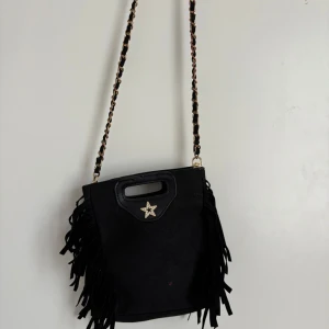 Svart axelväska med fransar och stjärndetalj - Cool svart axelväska med långa fransar på sidorna och en kedja med svarta detaljer som axelrem. Väskan har ett handtag upptill och en glittrig stjärna framtill. Materialet ser ut som mocka och ger en mjuk vibe. Perfekt för dig som gillar statement-accessoarer.
