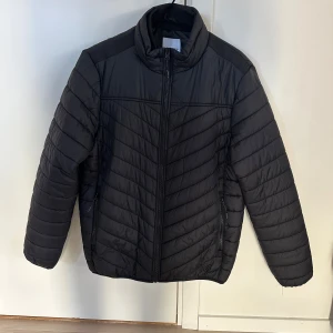 Svart dunjacka Lindbergh, herr M - Svart dunjacka från Lindbergh i relaxed fit, storlek M. Jackan har quiltad design med horisontella och snedställda sömmar, hög krage och dragkedja framtill. Köpt 2024, mycket fint skick. 