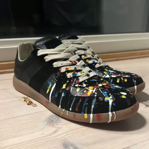 Svarta sneakers med färgstänk - Unika svarta sneakers med coola färgstänk i rött, gult, blått och vitt över hela ovandelen. Skorna har vita snören och en beige sula. Tillverkade i skinn med rund tå och platt sula. Perfekt för dig som vill sticka ut med din stil.