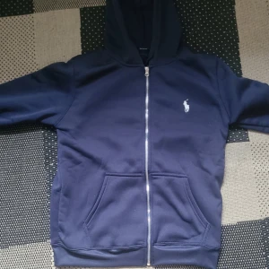Marinblå hoodie från Polo Ralph Lauren - Säljer en marinblå hoodie från Polo Ralph Lauren med dragkedja och huva. Tröjan har två stora fickor framtill och den klassiska vita Polo-loggan på bröstet. Perfekt för chill dagar och enkel att matcha med jeans eller joggers.
