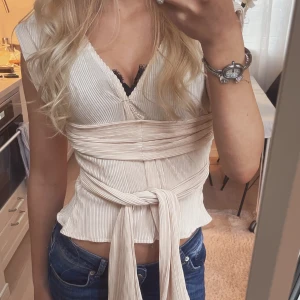 Plisserad omlottopp med knytband - Snygg ljusbeige omlottopp med plisserat tyg och breda knytband i midjan. Toppen har v-ringning och korta ärmar, perfekt att styla med jeans eller kjol. Materialet känns mjukt och följsamt, och knytet ger en cool och trendig look.