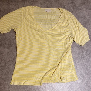 Gul och vit randig topp Holly & White - Superfin topp från Holly & White i gul och vit randigt. Modellen har v-ringning och korta ärmar, perfekt för en fräsch och somrig look. Tunt och mjukt material som känns skönt mot huden.