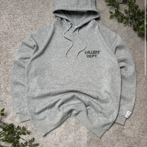 Grå hoodie från Gallery Dept. - Snygg grå hoodie från Gallery Dept. med svart tryck fram och stort tryck på ryggen. Klassisk huva med snörning och ribbade muddar. Mjuk och skön i bomullsmix, perfekt för chill dagar. Liten vit etikett på ärmen med text.