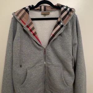 Grå hoodie från Burberry  - Grå hoodie från Burberry med klassisk rutig huva i beige, svart och röd. Tröjan har dragkedja framtill, snörning vid huvan och två fickor. Broderad Burberry-logga på bröstet. Tillverkad i mjuk bomull med fleecefoder för extra komfort. Säljer den eftersom jag använder inte den längre storlek M men passar även S pris 800 kan diskuteras vid snabb affär