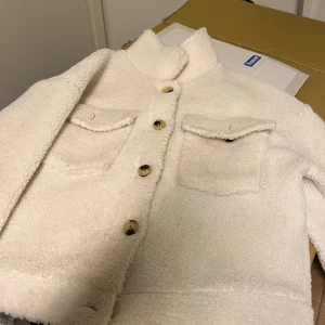 Vit teddyjacka med fickor och knappar - Säljer en snygg vit teddyjacka med två stora bröstfickor och coola knappar framtill. Jackan har en rak passform och hög krage, perfekt för att hålla värmen. Materialet är mjukt och fluffigt, vilket ger en trendig look.