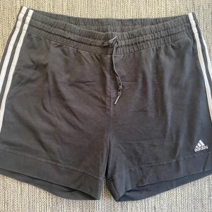 Svarta Adidas shorts med snörning XL - Svarta shorts från Adidas i storlek XL med klassiska vita ränder längs sidorna och snörning i midjan. Mjukt bomullsmaterial och elastisk midja för skön passform. Perfekta för träning eller chill dagar.      Mått Midjan: ca 46 cm Längd från grenen och ner: ca 11 cm  93% bomull och 7% spandex  Kommer från ett djur och rökfritt hem.
