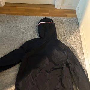 Svart vindjacka från Moncler med huva - Svart vindjacka från Moncler med klassisk logga på ärmen och huva med vit och röd rand. Jackan har dragkedja framtill och är tillverkad i lätt polyester, perfekt för blåsiga dagar. Snygg och stilren design som passar till det mesta.