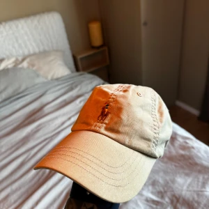 Beige keps från Polo Ralph Lauren - Snygg beige keps från Polo Ralph Lauren med klassisk broderad logga framtill och justerbart spänne bak i brunt läder. Kepsen är i bomull och har en avslappnad, tidlös look som passar perfekt till streetwear eller casual outfits.