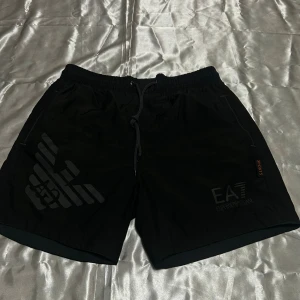 Svarta EA7 Emporio Armani shorts - Svarta shorts från EA7 Emporio Armani med sportig look, elastisk midja och snörning. Stora logotryck på ena benet och diskret 'SPORTS'-detalj. Perfekta för träning eller chill dagar. Lätta och bekväma med fickor på sidorna och bak.