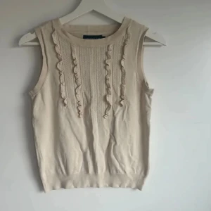 Beige ärmlös topp från Stockh LM - Supersöt beige linnetopp från Stockh LM med ribbad struktur och dekorativa volanger framtill. Rund halsringning och ärmlös design gör den perfekt för varma dagar. Materialet är mjukt och stickat, vilket ger en skön känsla och snygg passform.