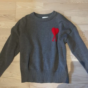 Grå stickad tröja från Ami Paris - Snygg grå stickad tröja från Ami Paris med rund hals och ribbade muddar. På bröstet finns Ami:s ikoniska röda hjärta med bokstaven A. Perfekt för dig som vill ha en stilren men ändå unik look.