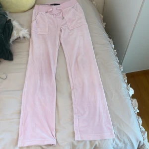 Ljusrosa mjukisbyxor Juicy Couture XS - Supermjuka ljusrosa mjukisbyxor från Juicy Couture med bred resår i midjan och snörning. Byxorna har raka ben, stora fickor framtill och klassisk logga broderad på ena fickan. Perfekta för en chill och trendig look.