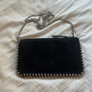 Svart axelväska med kedja och nitar - Snygg svart axelväska i mocka med silverfärgad kedja och coola nitar längs kanterna. Väskan har dragkedja upptill och flera fack inuti för smidig organisering. Perfekt för dig som gillar en edgy och stilren look.