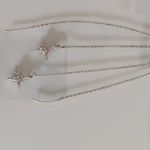 Silvriga örhängen med stjärnhänge - Långa, tunna örhängen i silverfärg med kedja och hängande stjärnor dekorerade med små glittrande stenar. Perfekta för dig som gillar unika och trendiga smycken med lite extra bling.
