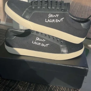 Svarta Saint Laurent sneakers strl 43 - Snygga svarta sneakers från Saint Laurent med vit handskriven logga på sidan. Skorna har svart canvas och skinnpaneler, samt vit platt sula i gummi. Klassisk låg modell med snörning och Saint Laurent Paris-detaljer på insidan och sulan. Perfekta för en stilren look.