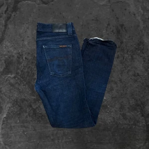Mörkblå jeans från Nudie Jeans - Snygga mörkblå jeans från Nudie Jeans med klassisk femficksdesign och raka ben. Jeansen har dragkedja i gylfen och subtila detaljer på bakfickorna. Tillverkade i slitstarkt denimtyg som ger en tidlös look.