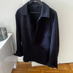 Mörkblå overshirt från COS - Stilren mörkblå overshirt från COS med klassisk krage och stora fickor framtill. Skjortan har knäppning hela vägen och är tillverkad i ett mjukt bomullstyg. Perfekt lager-på-lager-plagg med avslappnad passform och långa ärmar.