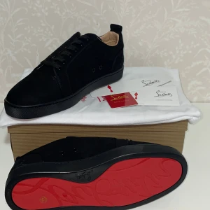 Svarta loubs - Snygga svarta sneakers från Christian Louboutin med klassisk röd sula och rund tå. Skorna har snörning och är tillverkade i mocka med svarta detaljer. Insidan är beige och sulan har Louboutins signatur. Perfekt för dig som gillar exklusiv streetstyle.