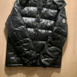 Svart Moncler pufferjacka herr - Säljer en svart pufferjacka från Moncler med glansig finish och klassisk logga på ärmen. Jackan har dragkedja framtill, fickor med knappar och quiltad design. Perfekt för dig som vill ha en snygg och varm jacka med streetkänsla.