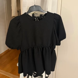 Svart blus med puffärm från Gina Tricot - Supersnygg svart blus från Gina Tricot med puffiga korta ärmar och volang nedtill. Toppen har öppen rygg med knytband både i midjan och nacken. Den är endast använd en gång.