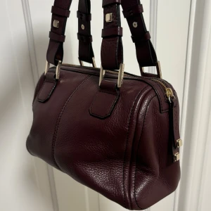 Speedy väska - Snygg vinröd handväska från Calvin Klein i äkta skinn med grov struktur. Väskan har guldfärgade detaljer, dubbla handtag med coola spännen och en dragkedja upptill. Utsmyckad med en unik tärningscharm som ger extra personlighet.