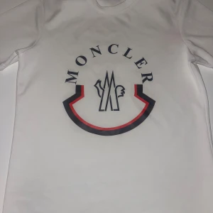 Vit Moncler t-shirt med stor logga - Snygg vit t-shirt från Moncler med stor tryckt logga i svart och rött på framsidan och mindre logga på ryggen. Klassisk rund hals och korta ärmar. Materialet känns som mjuk bomull, perfekt för en clean och stilren look. Passar xs-s