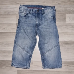 Blå jeansshorts med fickor - Säljer ett par blå jeansshorts med klassisk femficksdesign och knappstängning. Shortsen har en loose passform och är tillverkade i bomull. Snygga sömdetaljer på benen och bakfickor med knapp. Perfekta för dig som gillar avslappnad stil.