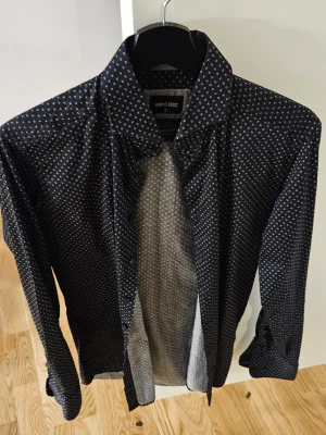 Svart prickig skjorta WAM Classic S - Svart skjorta från WAM Classic i storlek S med små vita prickar över hela tyget. Skräddarsydd passform och klassisk krage. Långärmad och med knappar framtill. Perfekt för dig som gillar stilrena detaljer och mönster.
