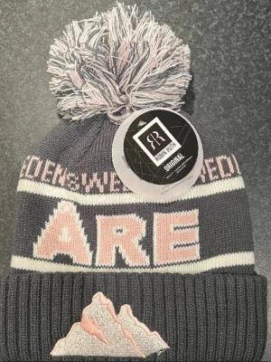 Mössa från Robin Ruth med Åre-tryck - Stickad mössa från Robin Ruth i mörkgrått med ljusrosa och vita detaljer. Stor tofs på toppen och broderat bergmotiv framtill. Texten 'ÅRE' i rosa och vitt. Helt ny skick och finns ändrats att köpa i butik på plats i Åre!
