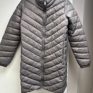 Grå/brun lång pufferjacka M - Säljer en grå/brun lång pufferjacka med dragkedja framtill och hög krage. Jackan har ett quiltat mönster och långa ärmar. Perfekt för kyligare dagar och enkel att styla till olika outfits.