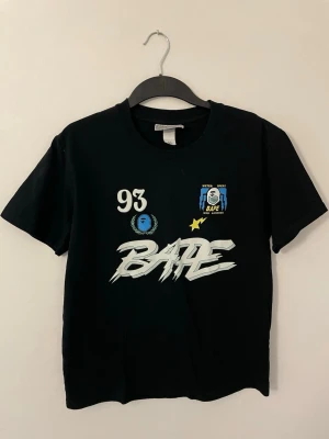 Svart BAPE t-shirt med tryck - Svart t-shirt från BAPE med stort grafiskt tryck på både fram- och baksidan. Framsidan har flera coola detaljer som siffran 93, stjärna och BAPE-logga i blått och vitt. Klassisk passform och rund hals. Perfekt för dig som gillar streetwear.