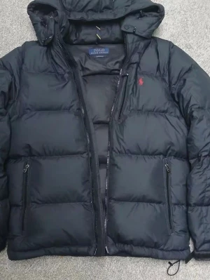 Svart pufferjacka från Polo Ralph Lauren - Svart pufferjacka från Polo Ralph Lauren med huva och dragkedja framtill. Jackan har två sidofickor med dragkedja och en röd Polo-logga på bröstet. Materialet är bomull och polyester, vilket gör den både varm och skön. Perfekt för kalla dagar.