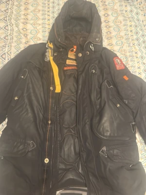 Svart Parajumpers pufferjacka med huva - Svart pufferjacka från Parajumpers med huva, coola patchar på ärmen och gul detaljrem vid kragen. Jackan har quiltad insida, flera fickor med dragkedja och knappar, samt orange och gula detaljer. Perfekt för dig som gillar streetstyle och vill ha något som sticker ut.