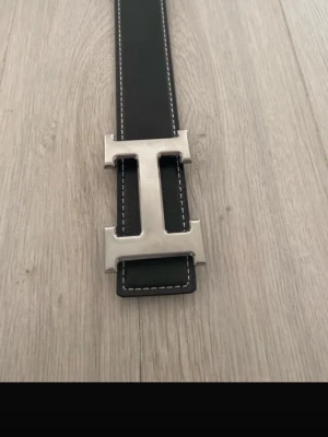 Svart skinnbälte med H-spänne - Snyggt svart bälte i skinn med ett stort, silvrigt H-format metallspänne. Bältet har vita sömmar längs kanterna som ger en schysst kontrast. Perfekt accessoar för att lyfta din outfit och ge en exklusiv vibe.