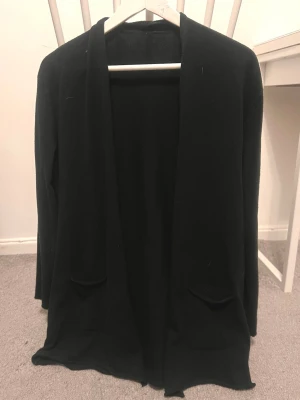 Svart lång cardigan jacka - Svart lång cardigan jacka med öppen front och två fickor framtill. Jackan har en enkel och stilren design med långärmade ärmar och mjukt material som ger en skön känsla. Perfekt för lager-på-lager och chill stil.