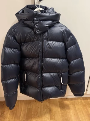Svart pufferjacka från J.Lindeberg - Säljer en svart pufferjacka från J.Lindeberg med huva och hög krage. Jackan har dragkedja framtill, två fickor med dragkedja och snygga detaljer med knappar vid halsen. Materialet är glansigt och fylligt, perfekt för kalla dagar.